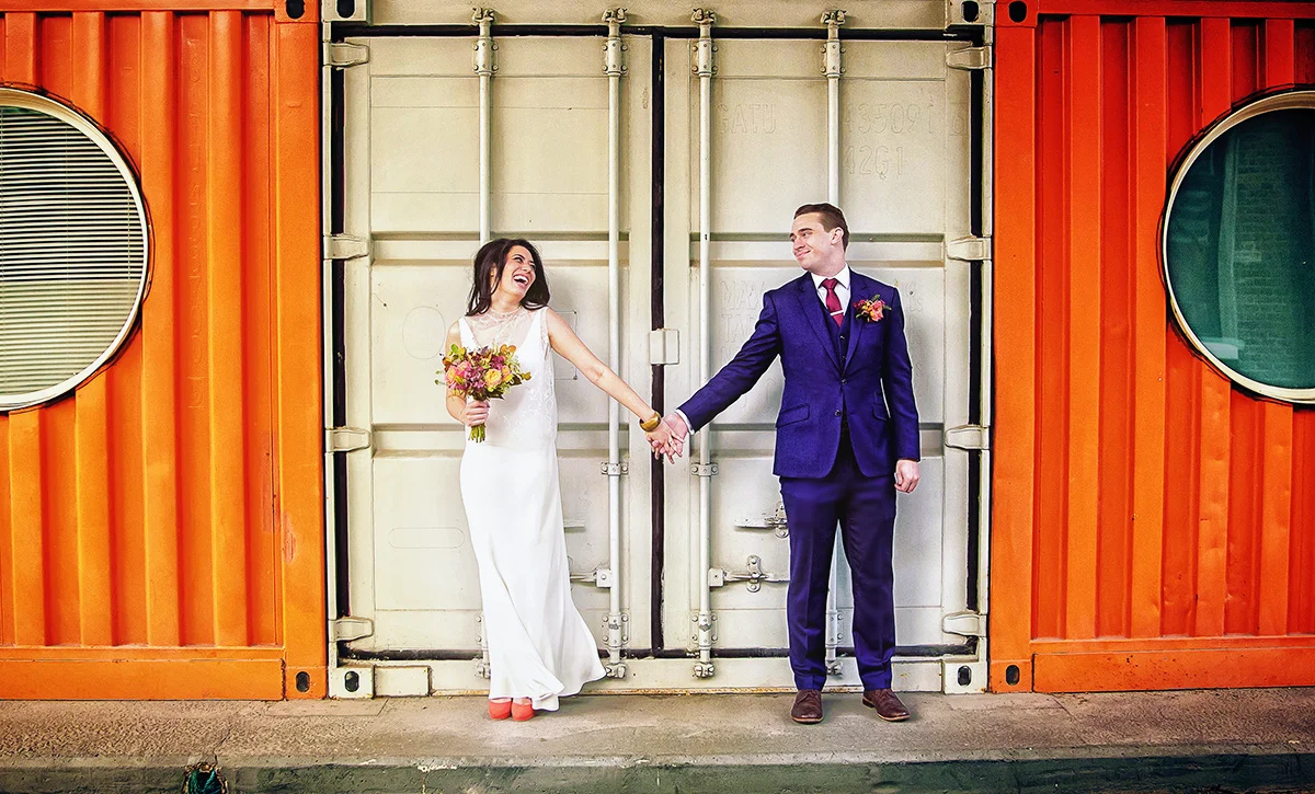 A wedding photographer duo capturing a love-filled moment between a bride and groom in front of an orange shipping container. (listing)