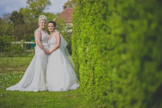 Two brides posing in front of a hedge.