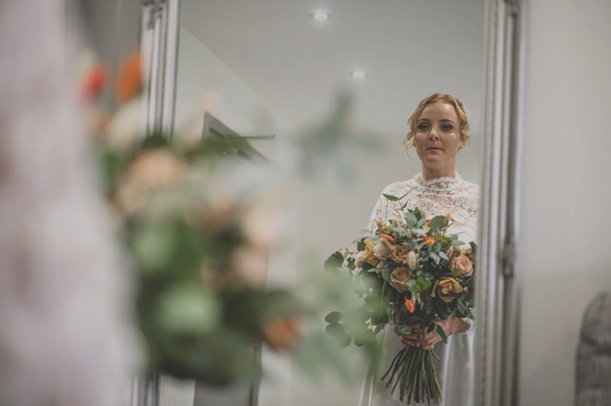 A bride is standing in front of a mirror holding her bouquet.