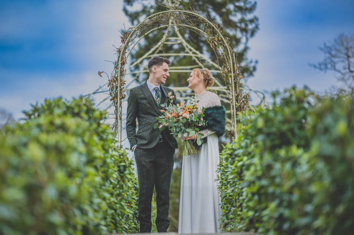 A bride and groom standing in front of an archway in a garden.