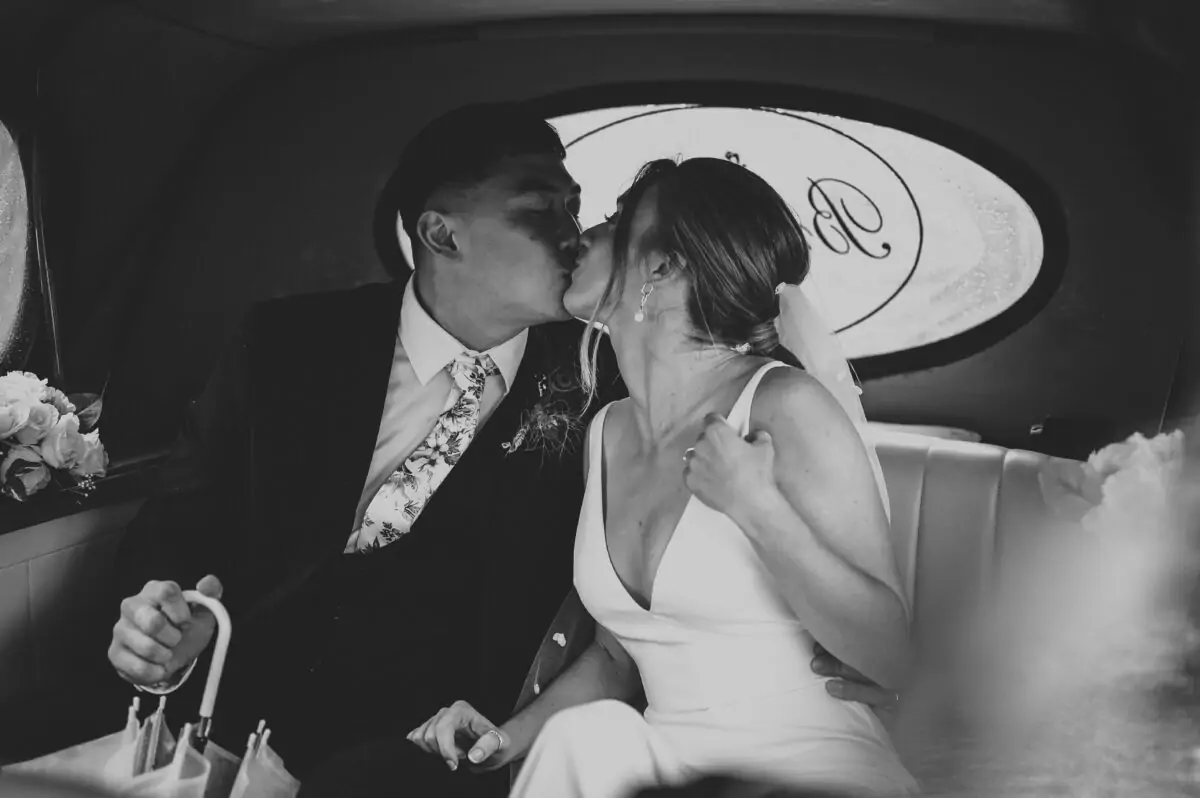 A bride and groom kiss in the back seat of a car.