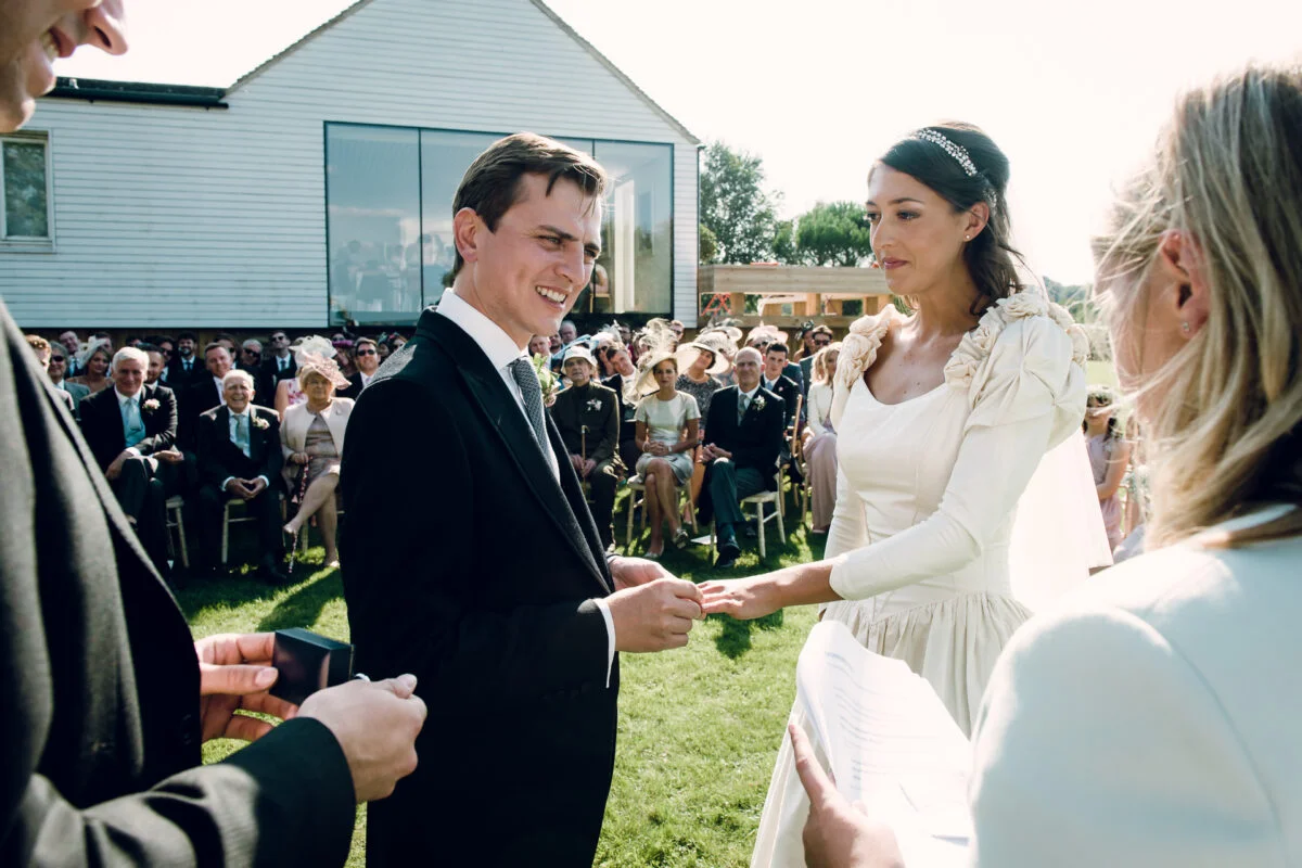 A smiling groom places a ring on the bride's finger during an outdoor ceremony on a bright sunny day with guests seated in the background.