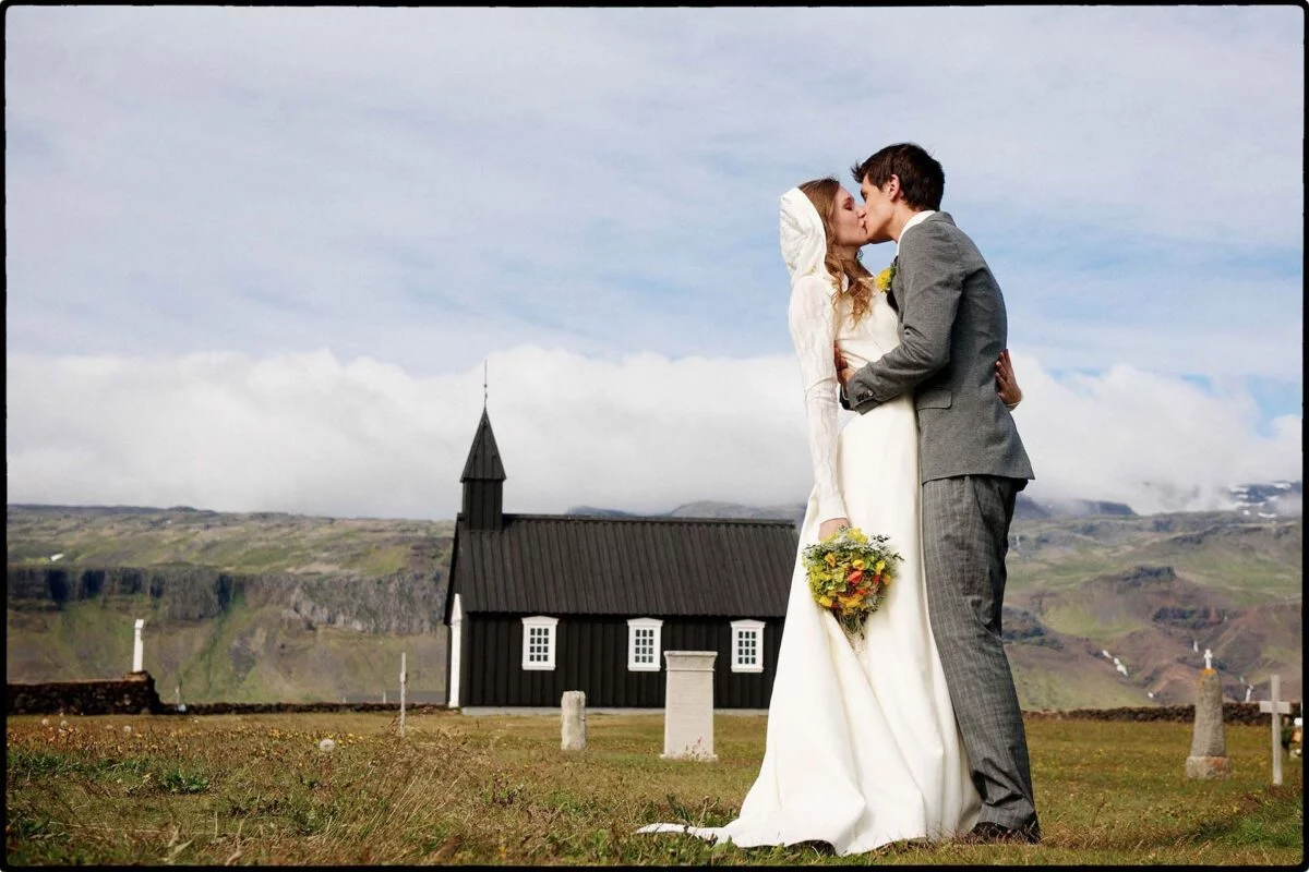 A newlywed couple shares a tender kiss outdoors in a dramatic mountainous landscape with a distinctive black church in the background.
