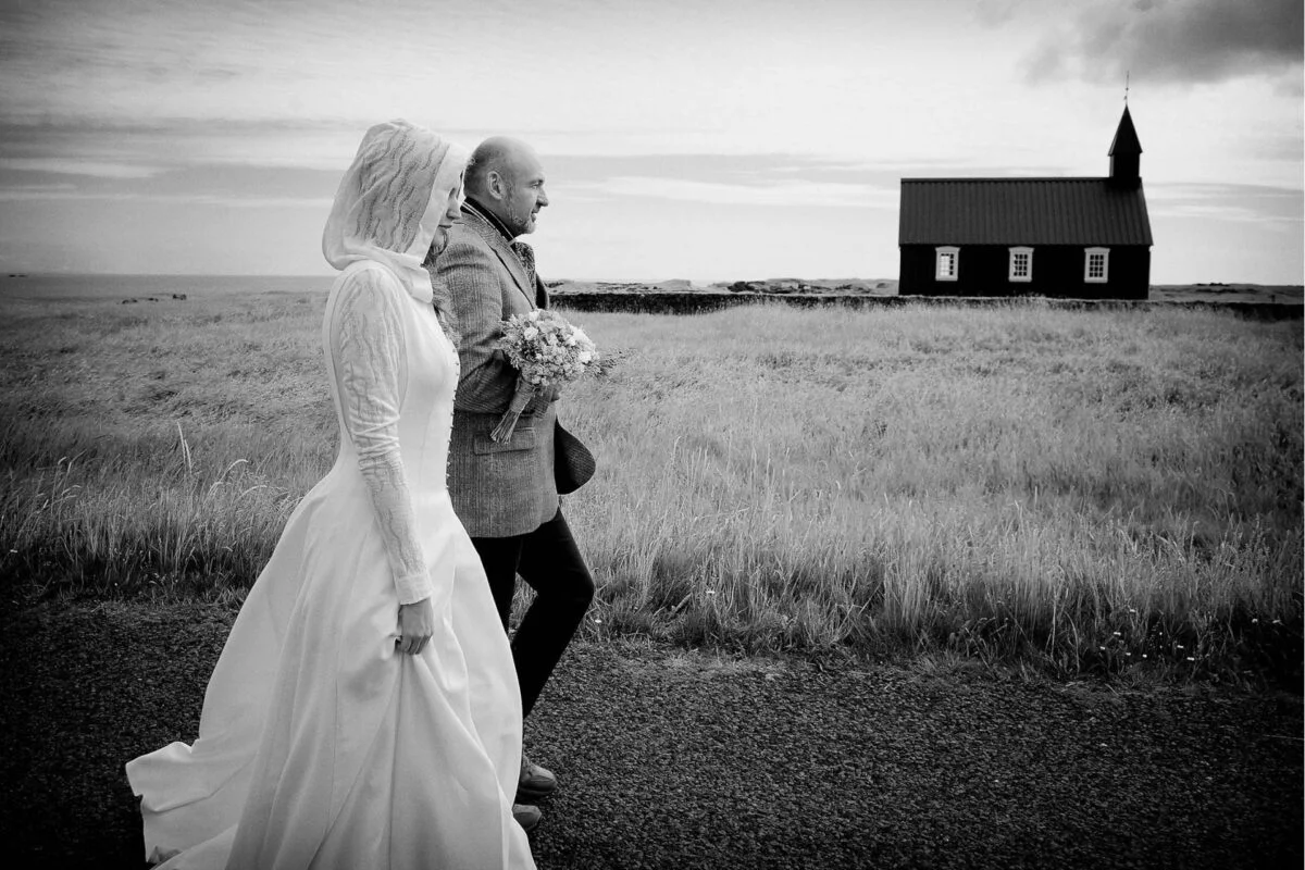 A bride and groom walking down a road with a church in the background.