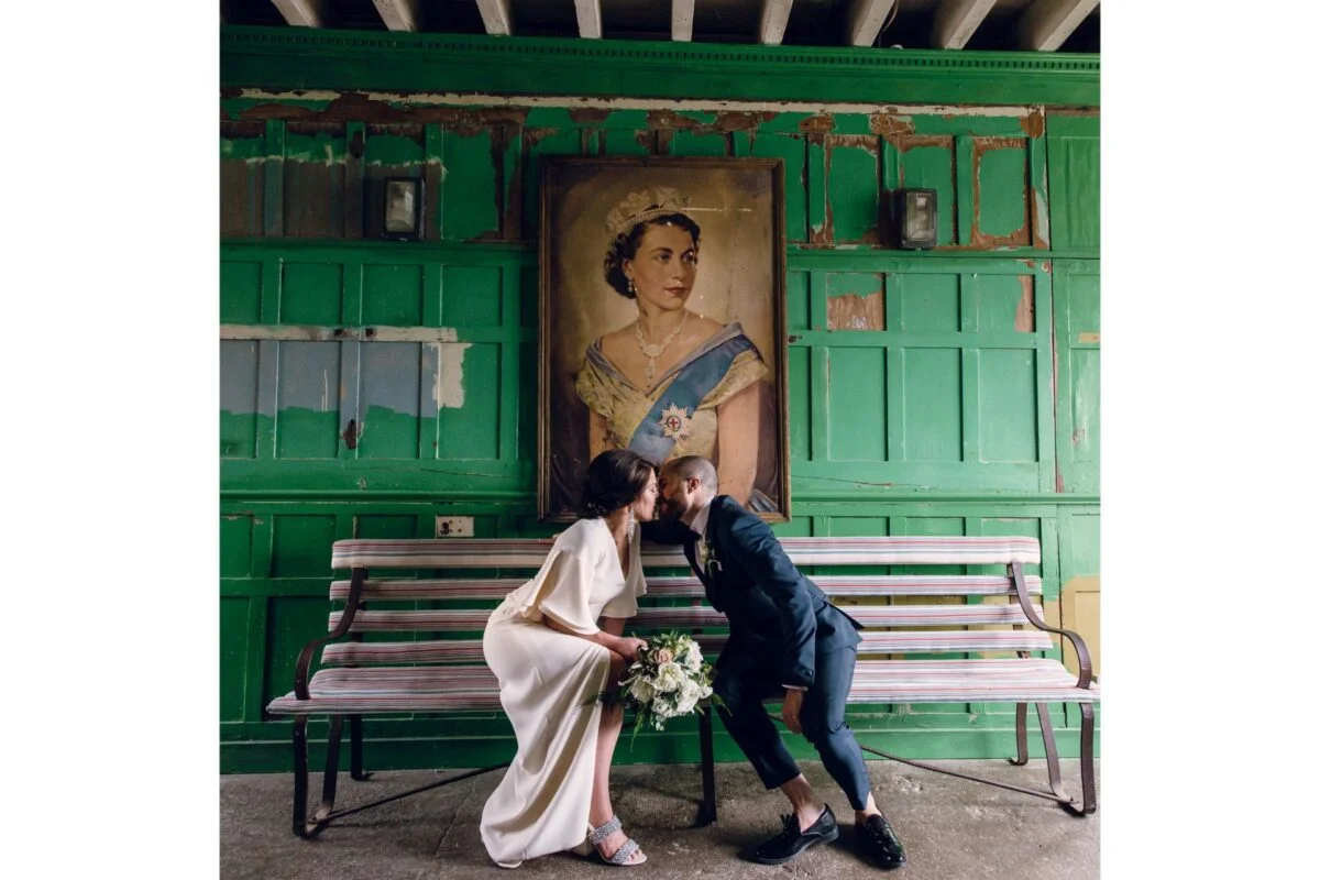 A bride and groom kissing on a bench in front of a painting.