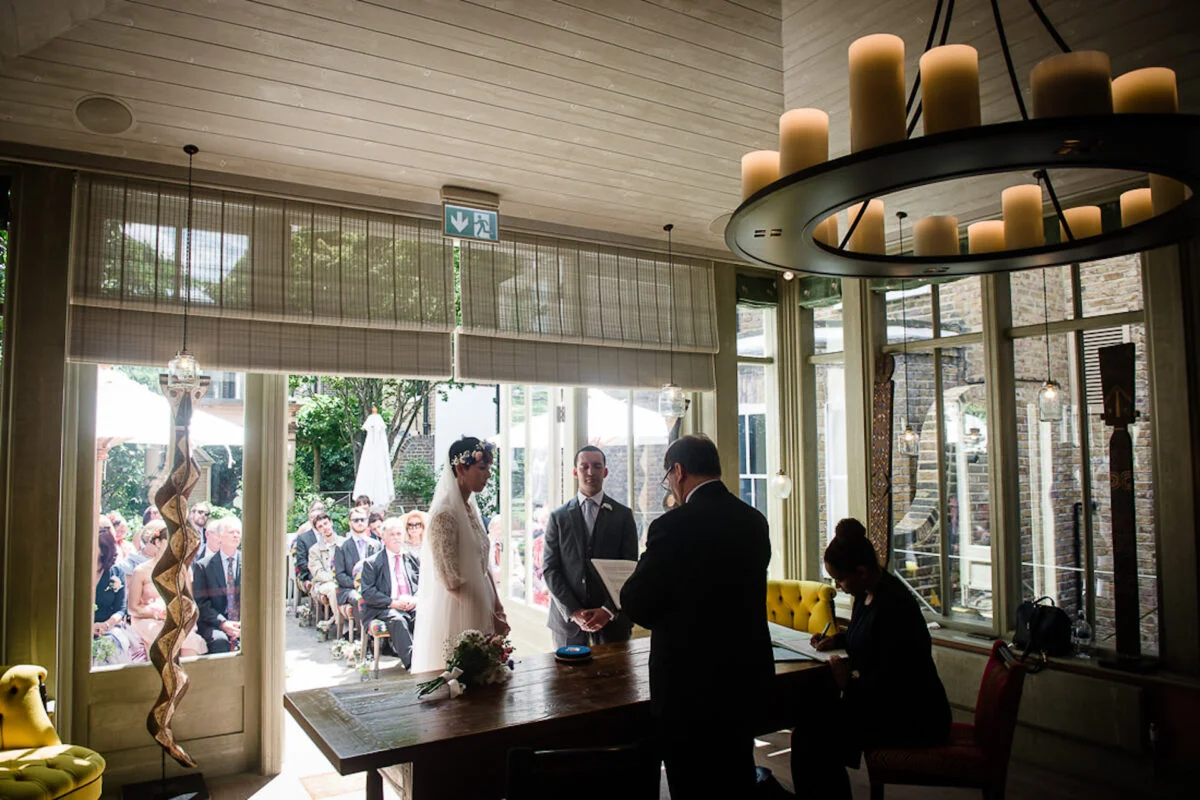 The bride and groom stand beside a wooden table during their light-filled civil ceremony while a registrar assists with signing the register.