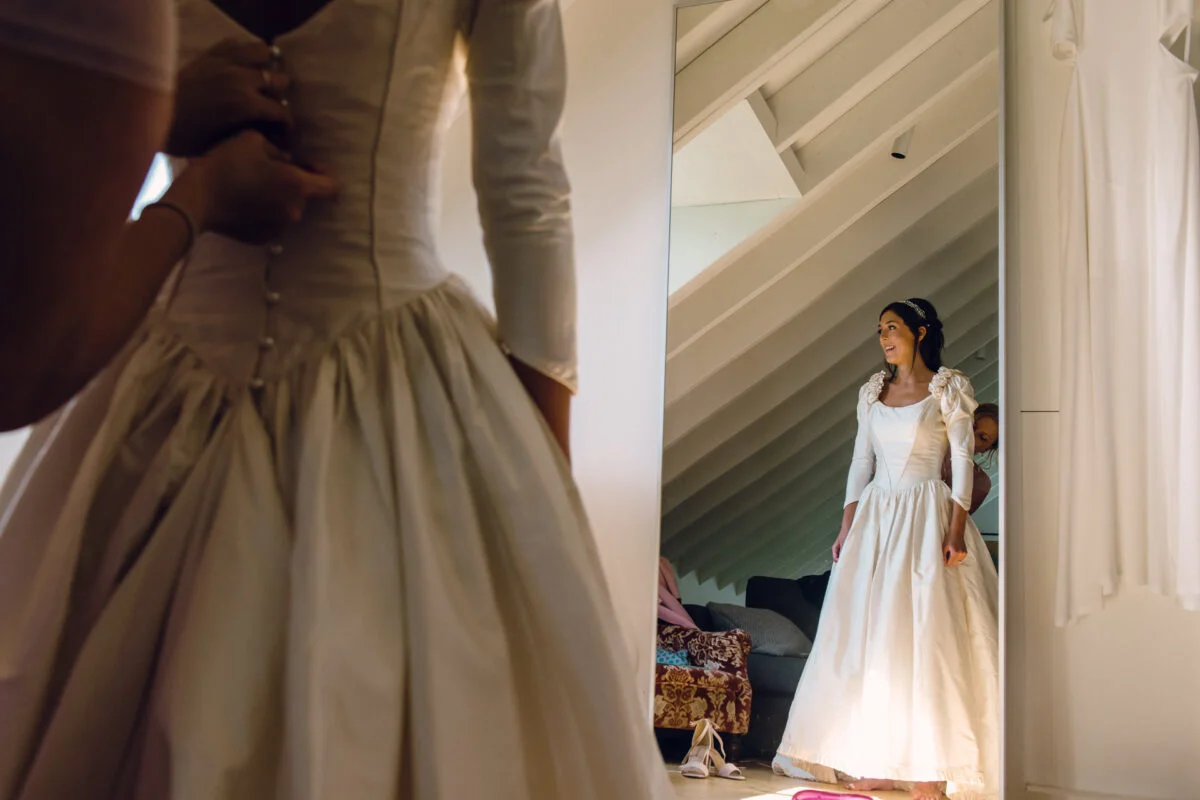A bride smiles as she looks into a full-length mirror while a bridesmaid buttons the back of her classic long-sleeved wedding dress.
