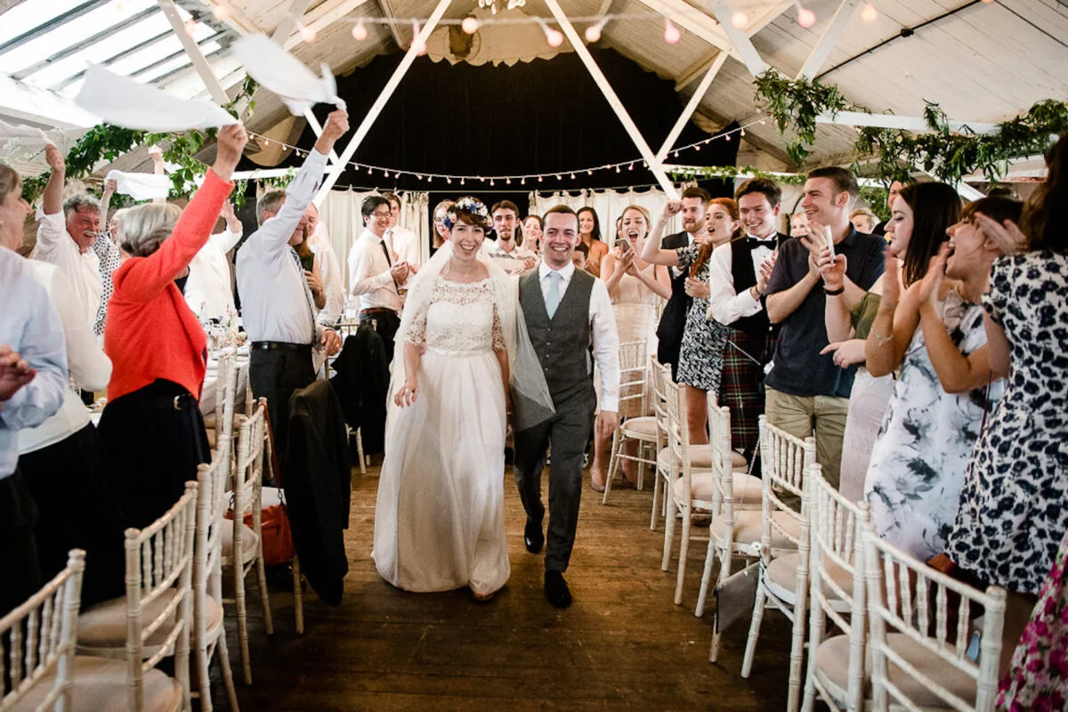 A smiling bride and groom walk hand in hand down the aisle of a rustic barn reception as guests cheer and wave white napkins.