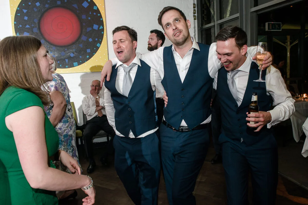 A lively group of groomsmen in navy waistcoats and suits dance together during an evening reception in an art gallery, holding drinks and laughing.
