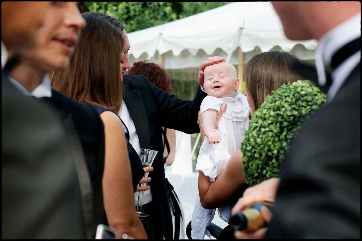 A smiling baby is held up and gently patted on the head by a guest among a crowd of people mingling outside near a white marquee.