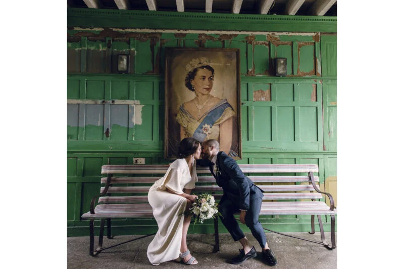 A bride and groom kissing on a bench in front of a painting.