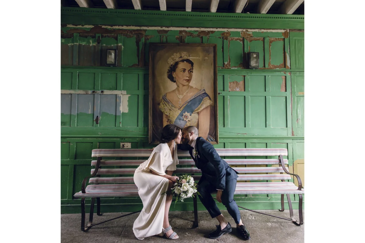 A bride and groom kissing on a bench in front of a painting.