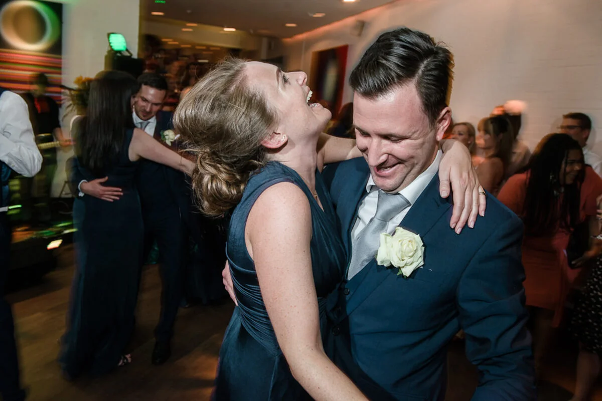 A joyful couple shares a moment of laughter while dancing together on a lively evening reception dance floor.