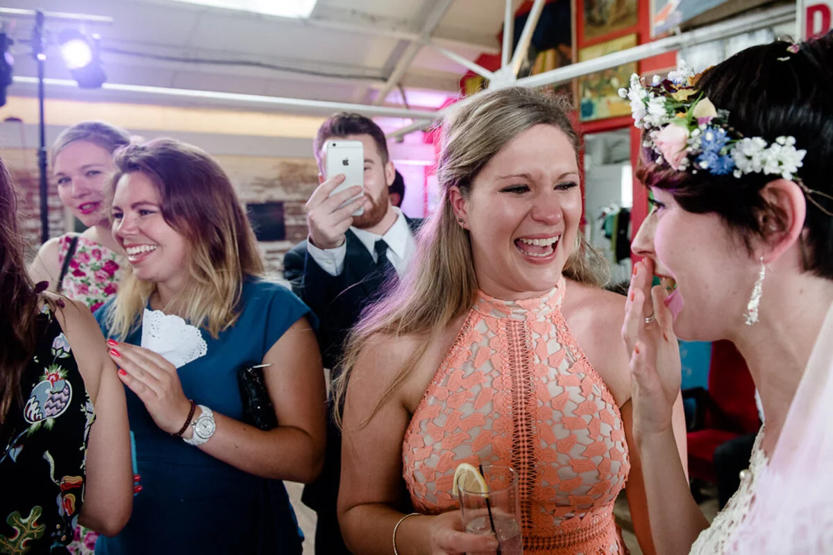 A group of joyful wedding guests laugh enthusiastically during the reception celebration, with one woman wearing a vibrant floral crown.