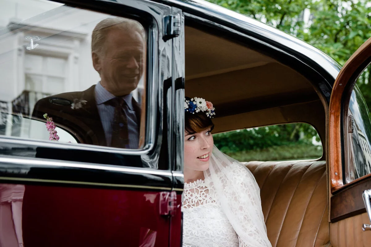 A smiling bride wearing a lace dress and flower crown looks out from the interior of a vintage wedding car while her father is reflected in the window.