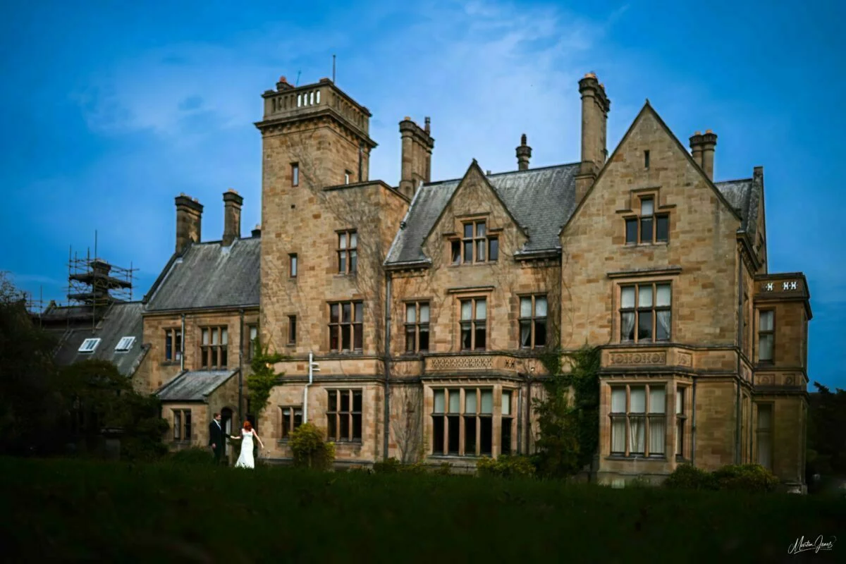 A bride and groom standing in front of a large mansion.