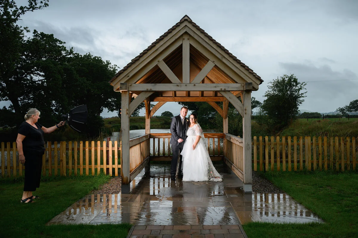A bride and groom standing under an umbrella in front of a gazebo.