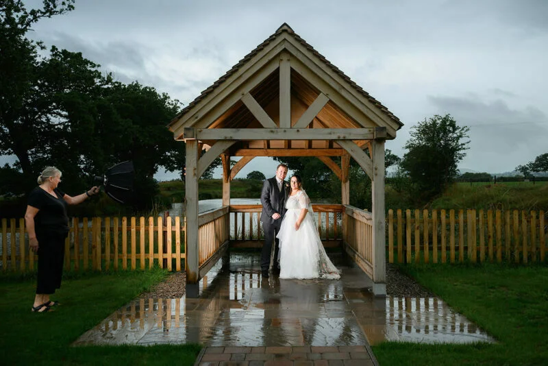 A bride and groom standing under an umbrella in front of a gazebo.