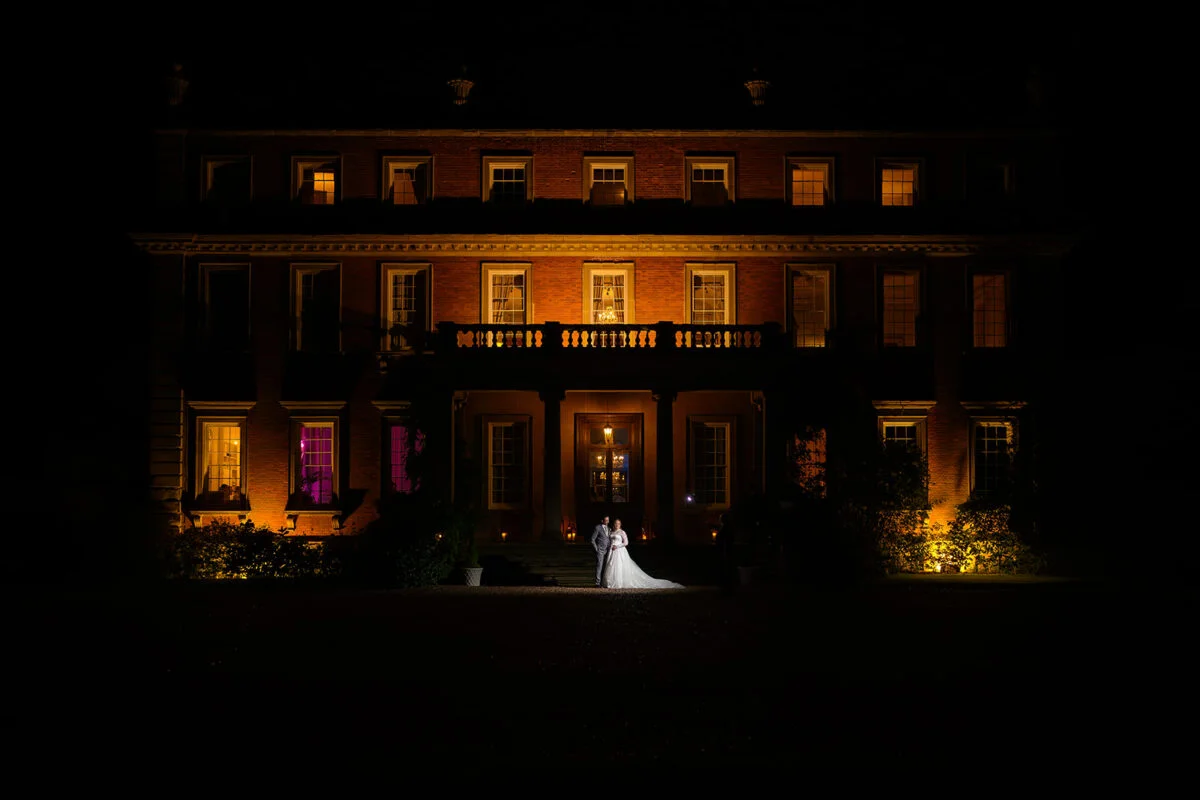 A bride and groom standing in front of a large mansion at night.