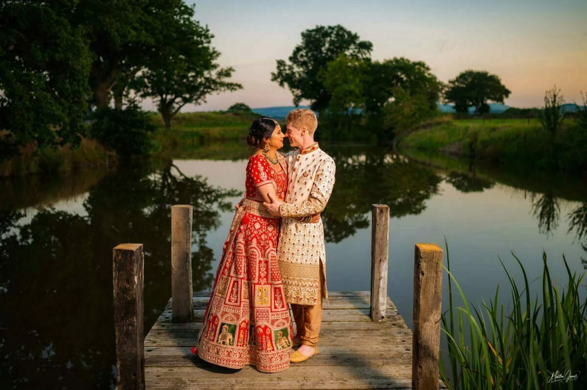A bride and groom standing on a dock near a pond.