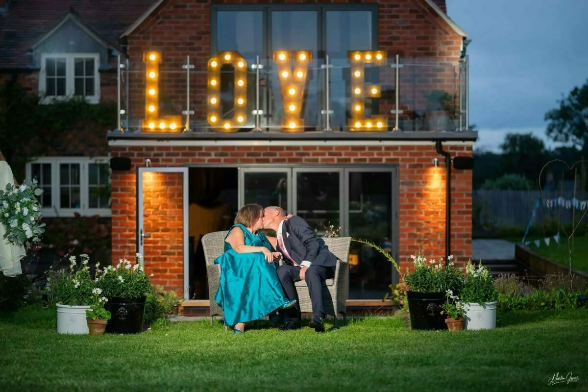 A bride and groom kissing in front of a lit up love sign.