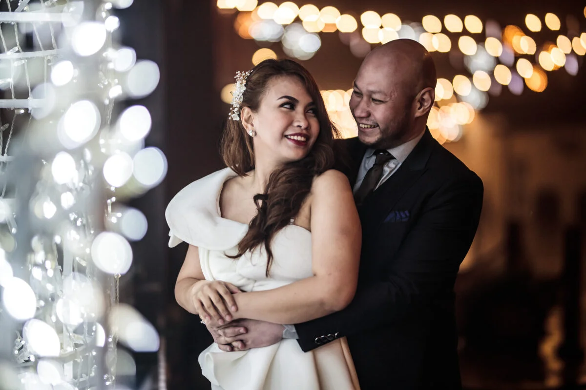 A bride and groom embracing in front of a string of lights.