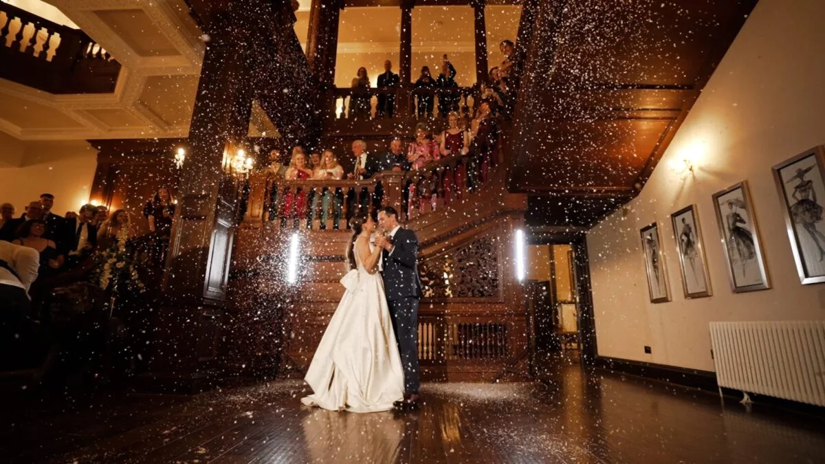 Newlyweds share their first dance in a grand panelled hall as a shower of white confetti cascades down around them while guests watch from the sweeping wooden staircase.