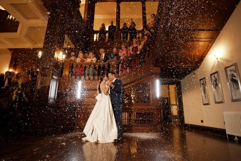 Newlyweds share their first dance in a grand panelled hall as a shower of white confetti cascades down around them while guests watch from the sweeping wooden staircase.