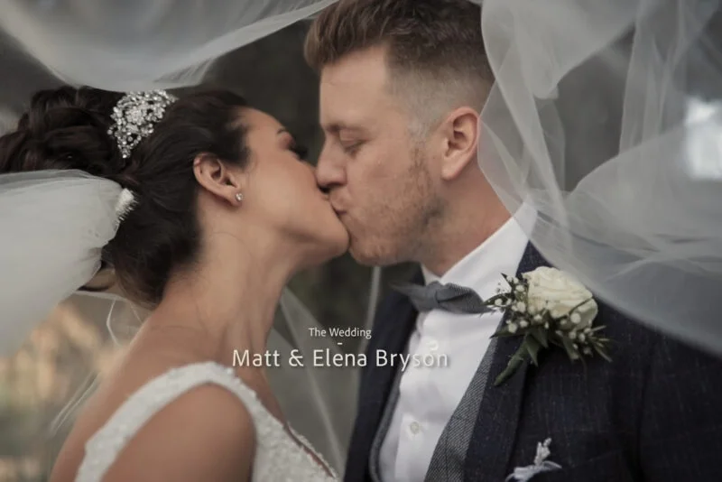 A newlywed couple share a tender kiss beneath a translucent veil framing their intimate portrait.