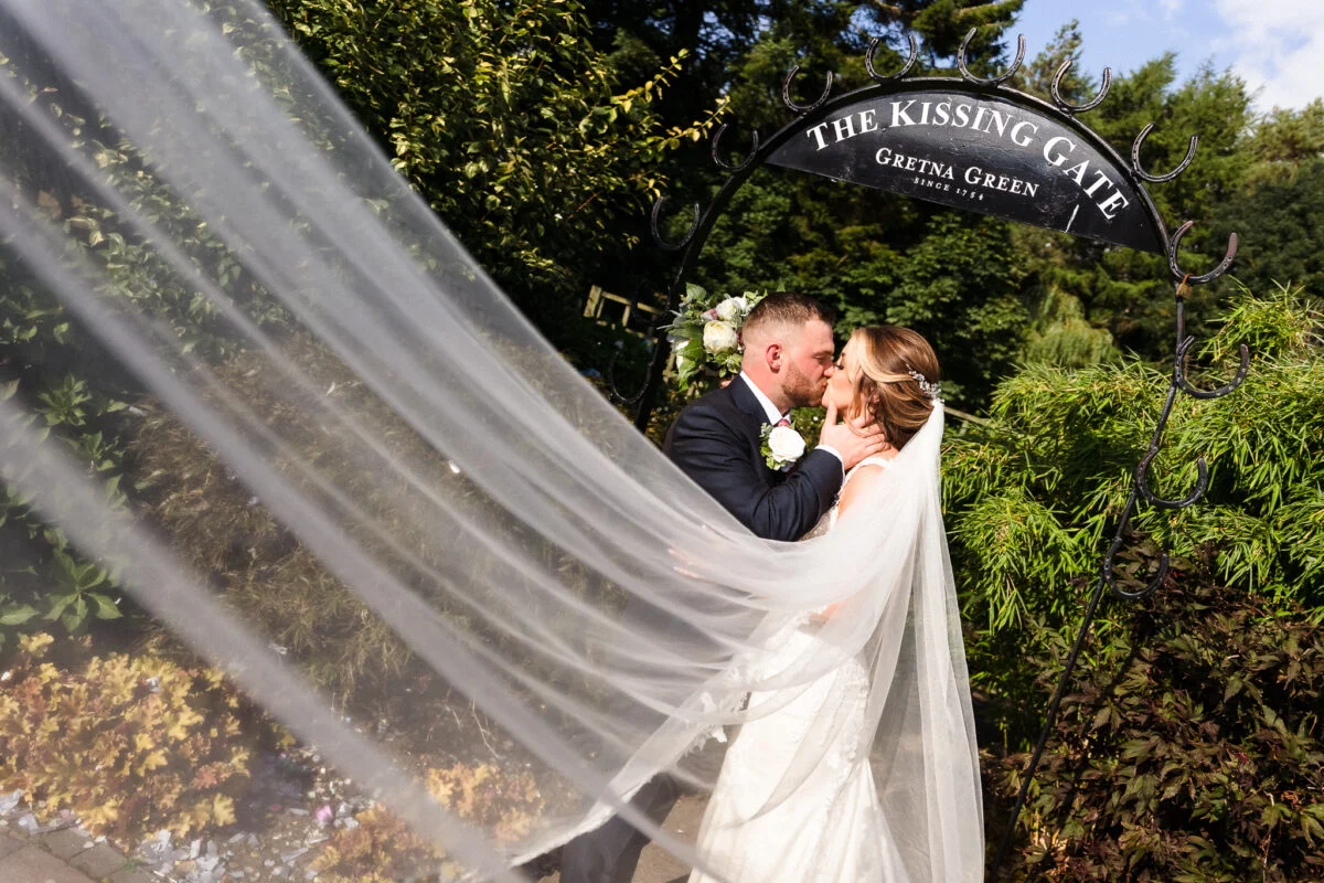 A bride and groom kiss under a veil in a garden.