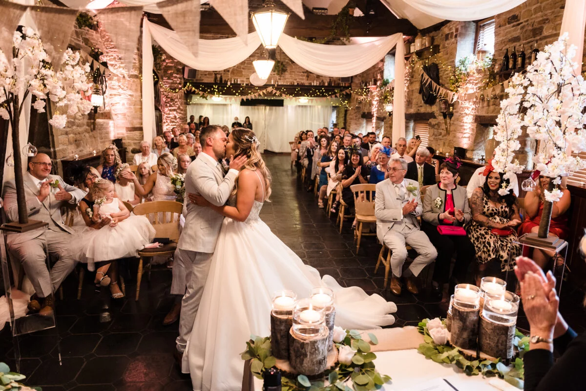 A bride and groom kiss during their wedding ceremony in a barn.