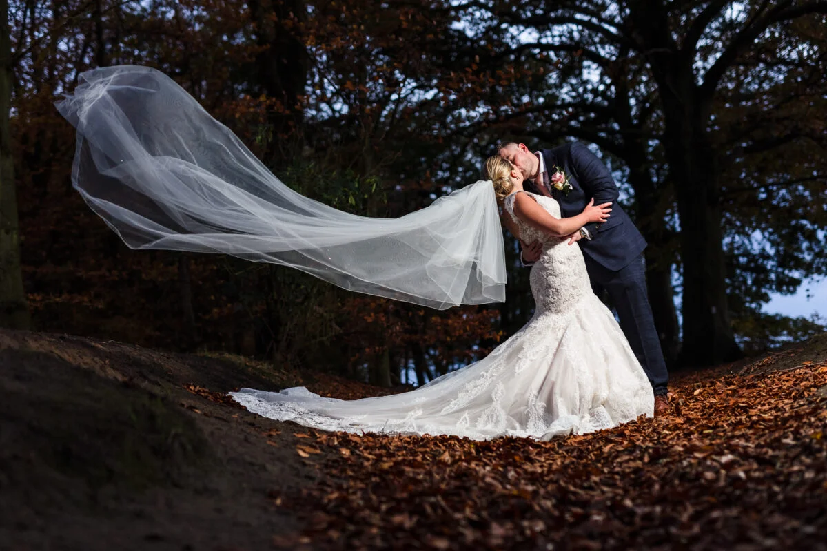A bride and groom kissing in the woods with their veil blowing in the wind.