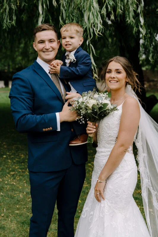 A man and woman in a wedding dress and a child holding a bouquet.