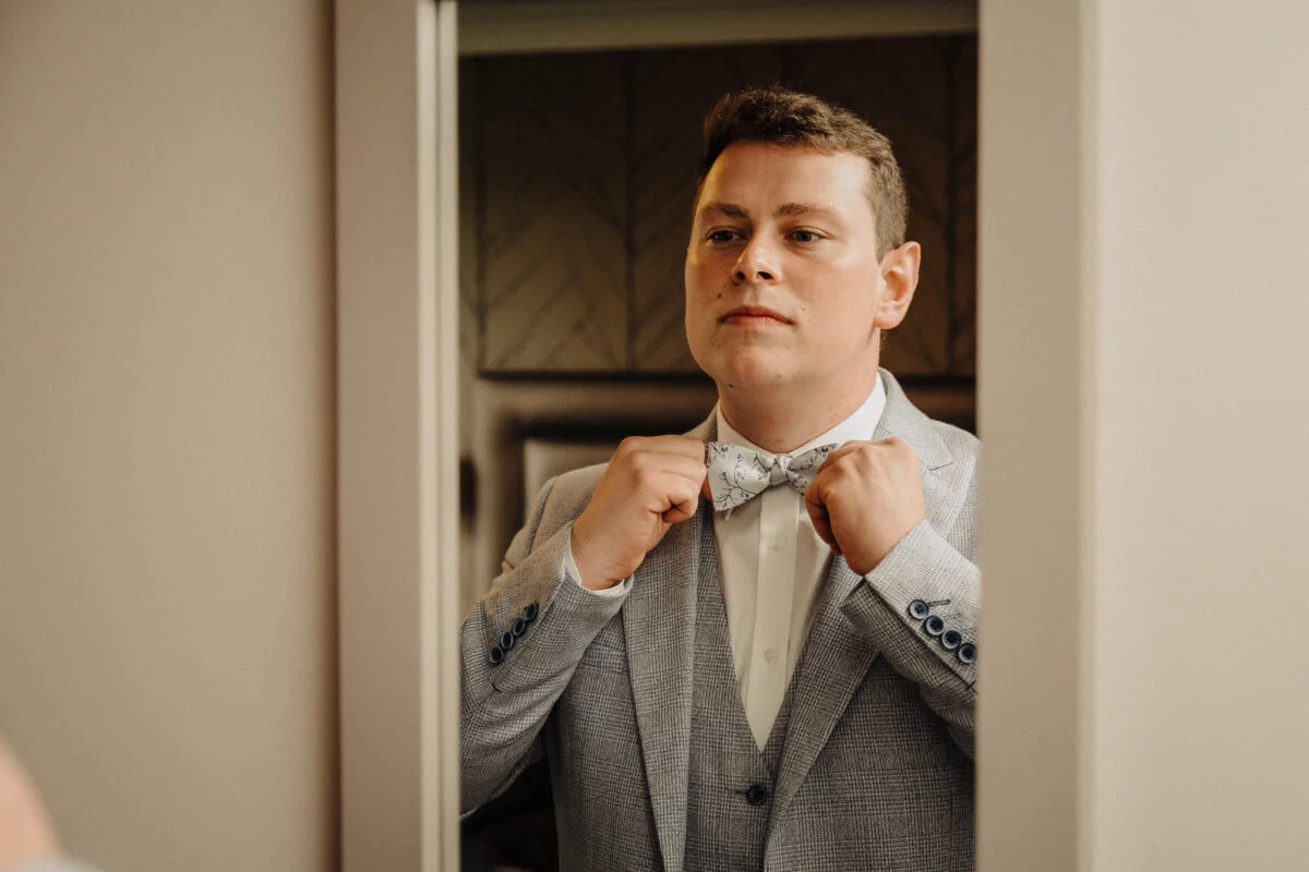 A man is adjusting his bow tie in a mirror.