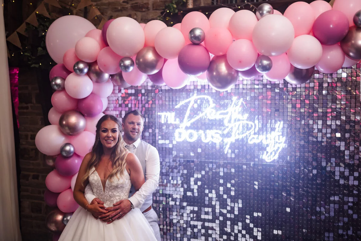 A bride and groom standing in front of a pink balloon arch.