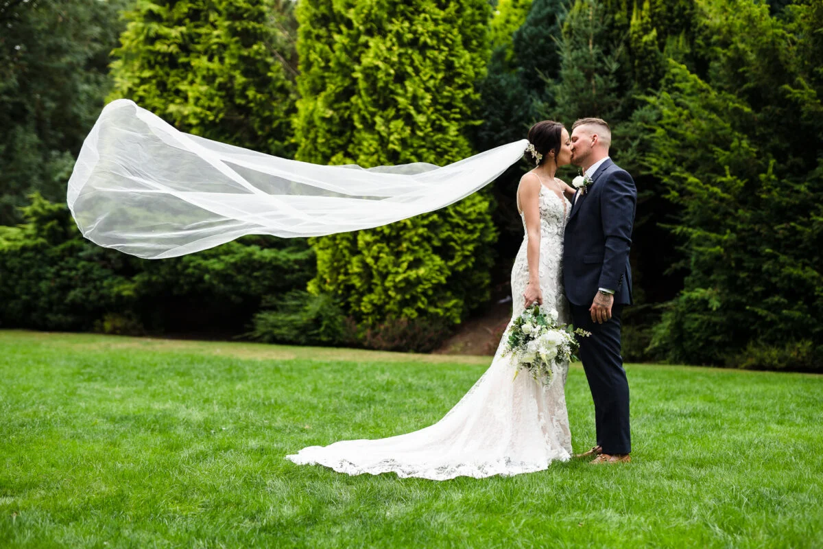 A bride and groom kissing in the grass with their veil blowing in the wind.