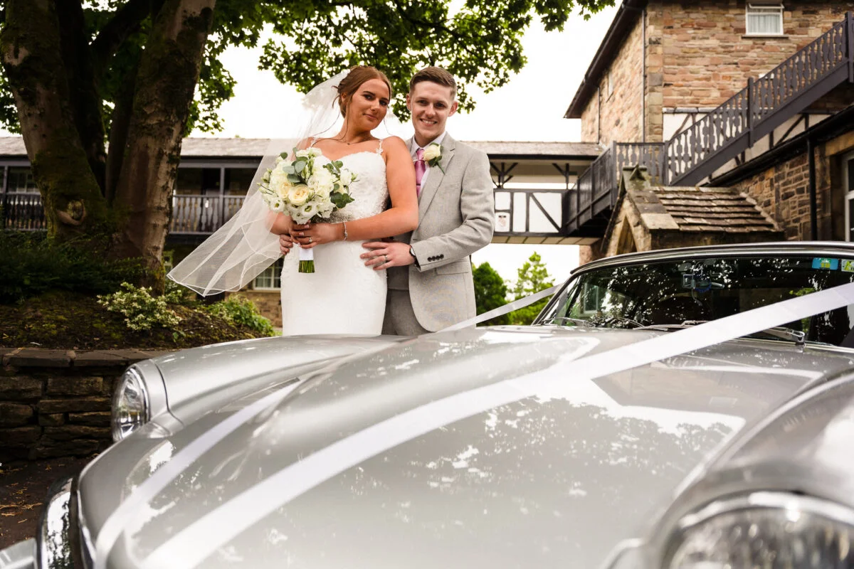 A bride and groom standing next to a silver sports car.