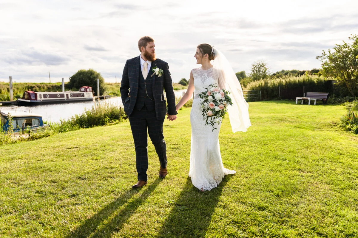 A bride and groom walking in the grass next to a boat.