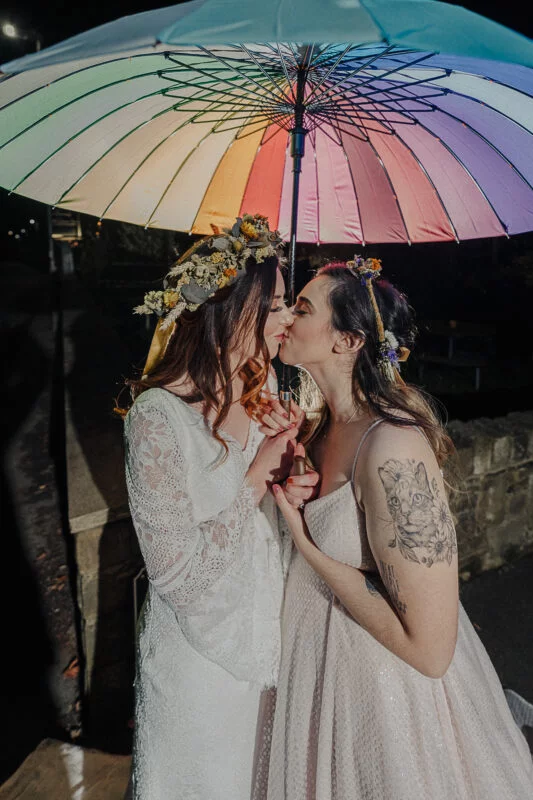 Two brides kiss under a rainbow umbrella.