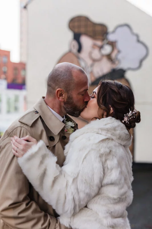 A bride and groom kiss in front of a mural.