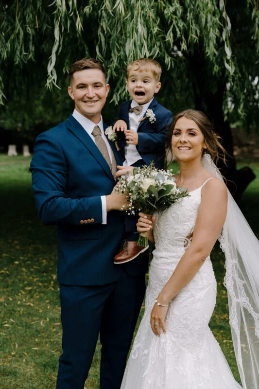 A bride and groom pose with their son under a willow tree.
