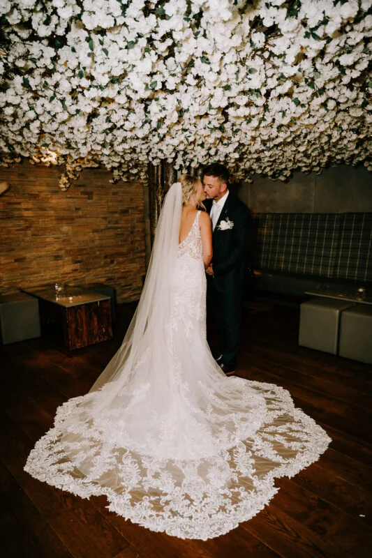 A bride and groom kiss under a flower covered ceiling.
