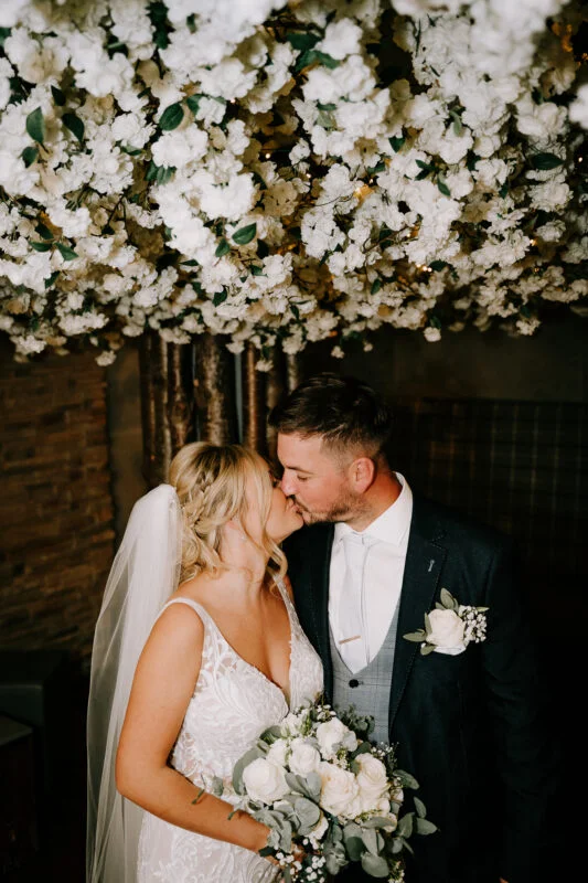 A bride and groom kiss under a flower covered ceiling.