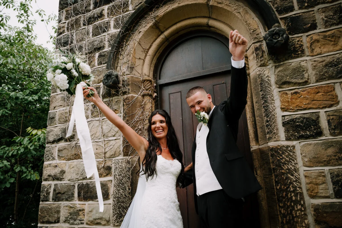 A bride and groom waving their arms in front of a stone building.