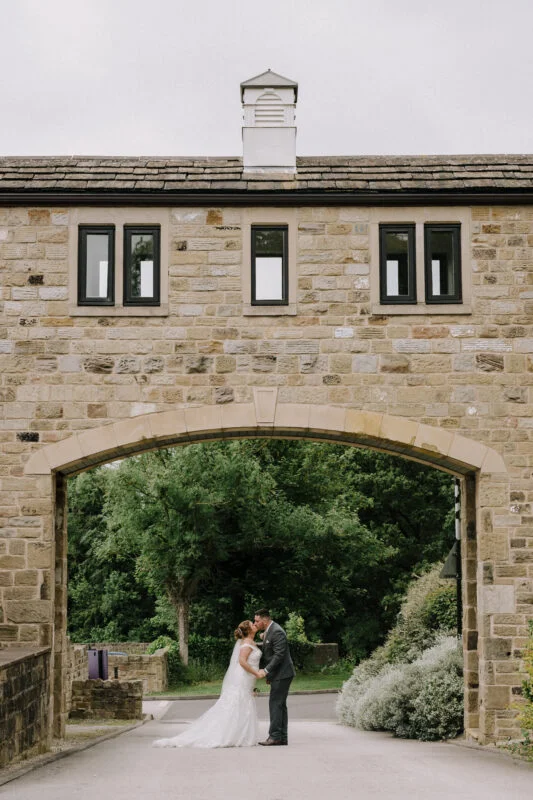 A bride and groom kissing in front of a stone archway.