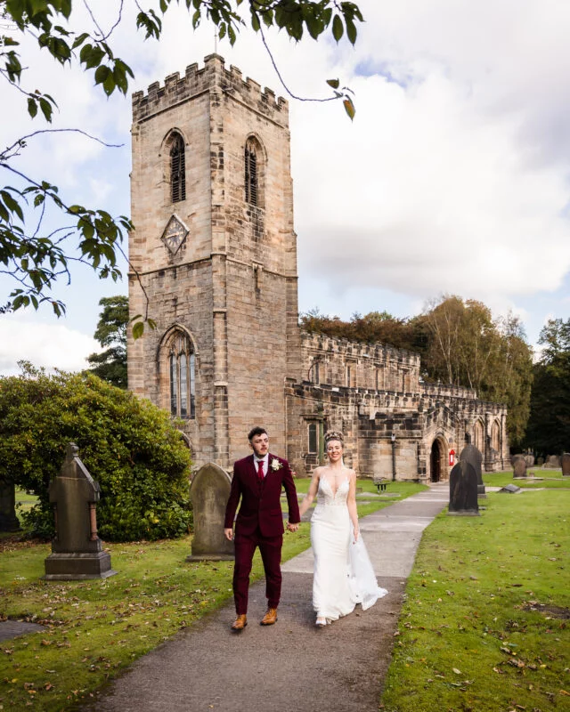 A bride and groom walking down a path in front of a church.