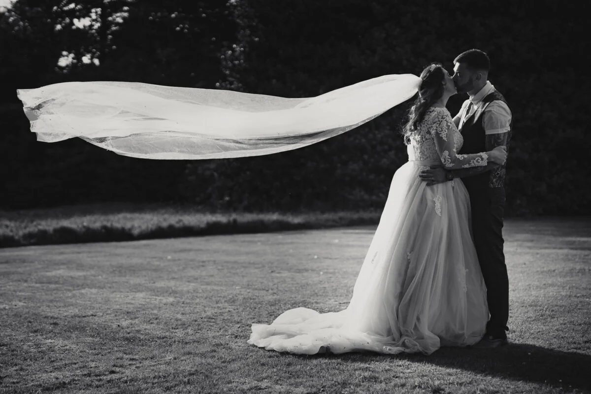 A bride and groom kissing in the grass with their veil blowing in the wind.