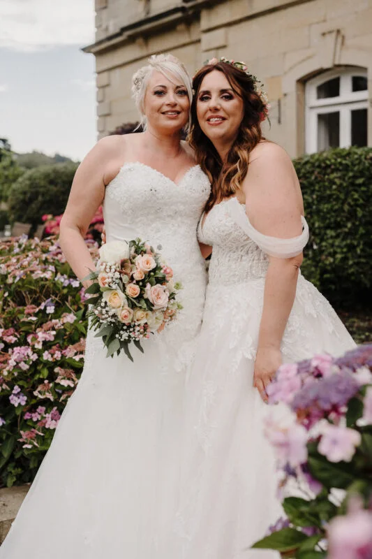 Two brides posing for a photo in front of flowers.