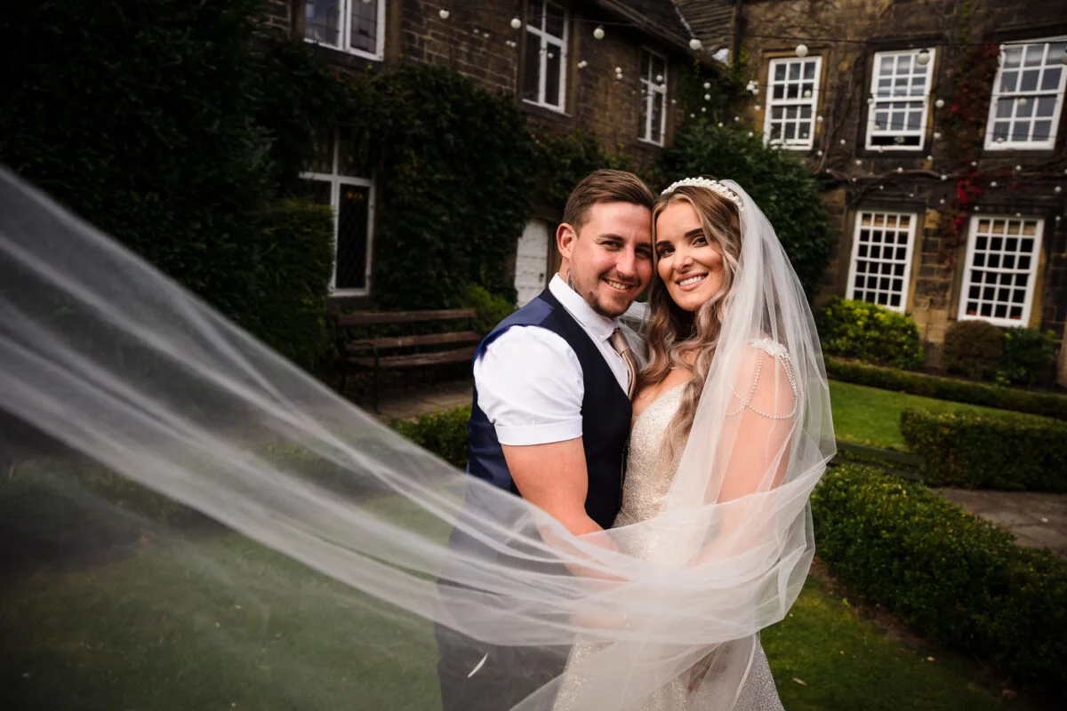A bride and groom posing in front of a building with a veil blowing in the wind.