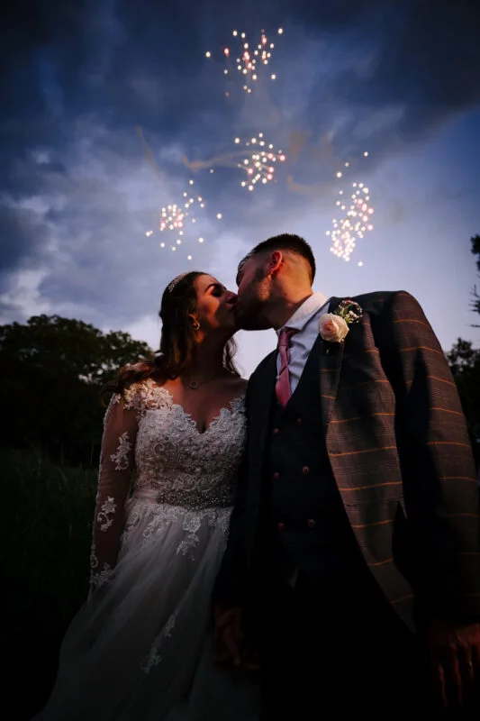 A bride and groom kiss in front of fireworks.
